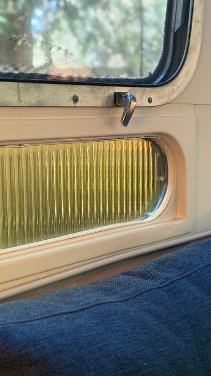 Airstream windows with a mesh screen and handle, and amber reeded window film viewed from inside, window above blue couch.